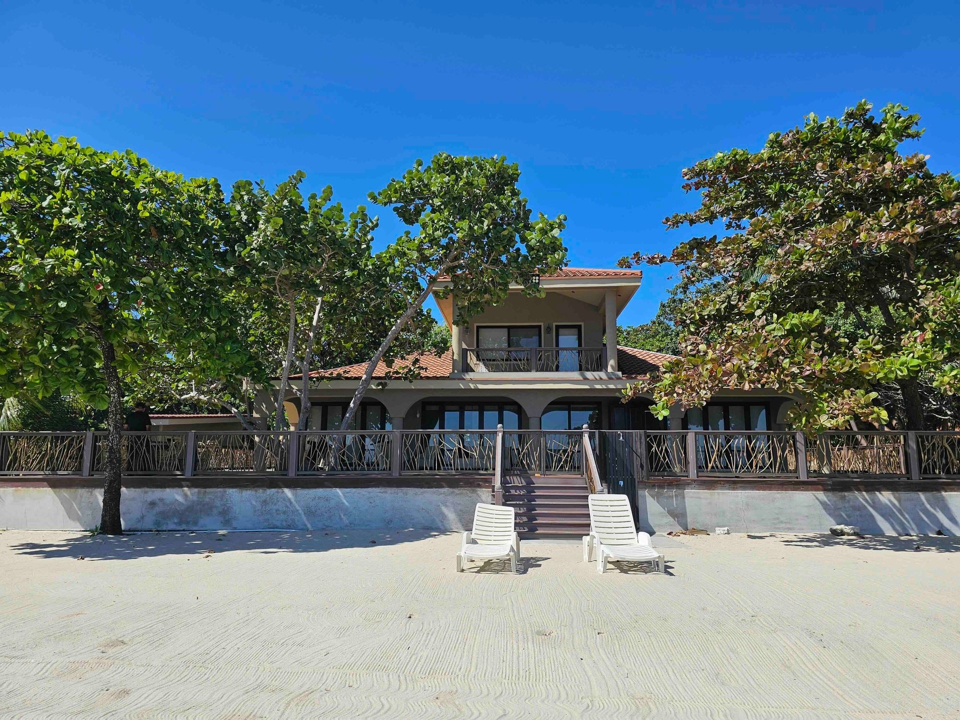 Beachfront house surrounded by trees with two white lounge chairs on sandy beach.