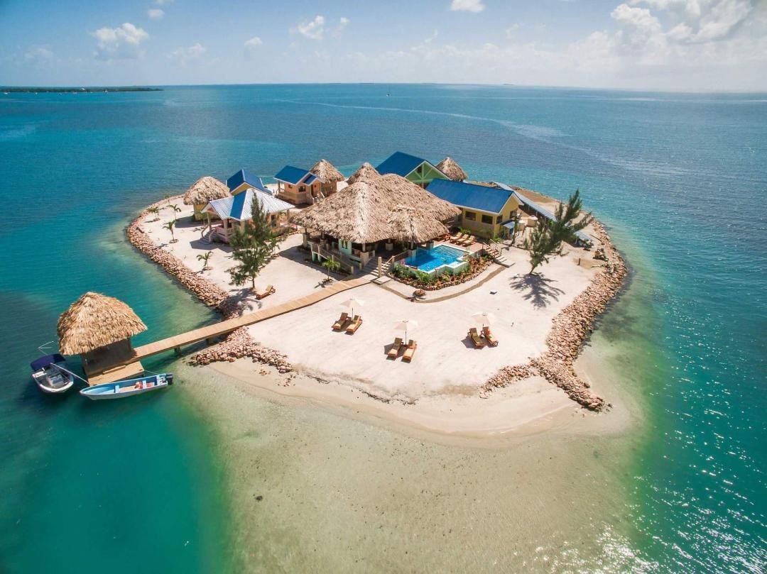 Aerial view of a tropical island with beach huts, pool, and boats on turquoise water.