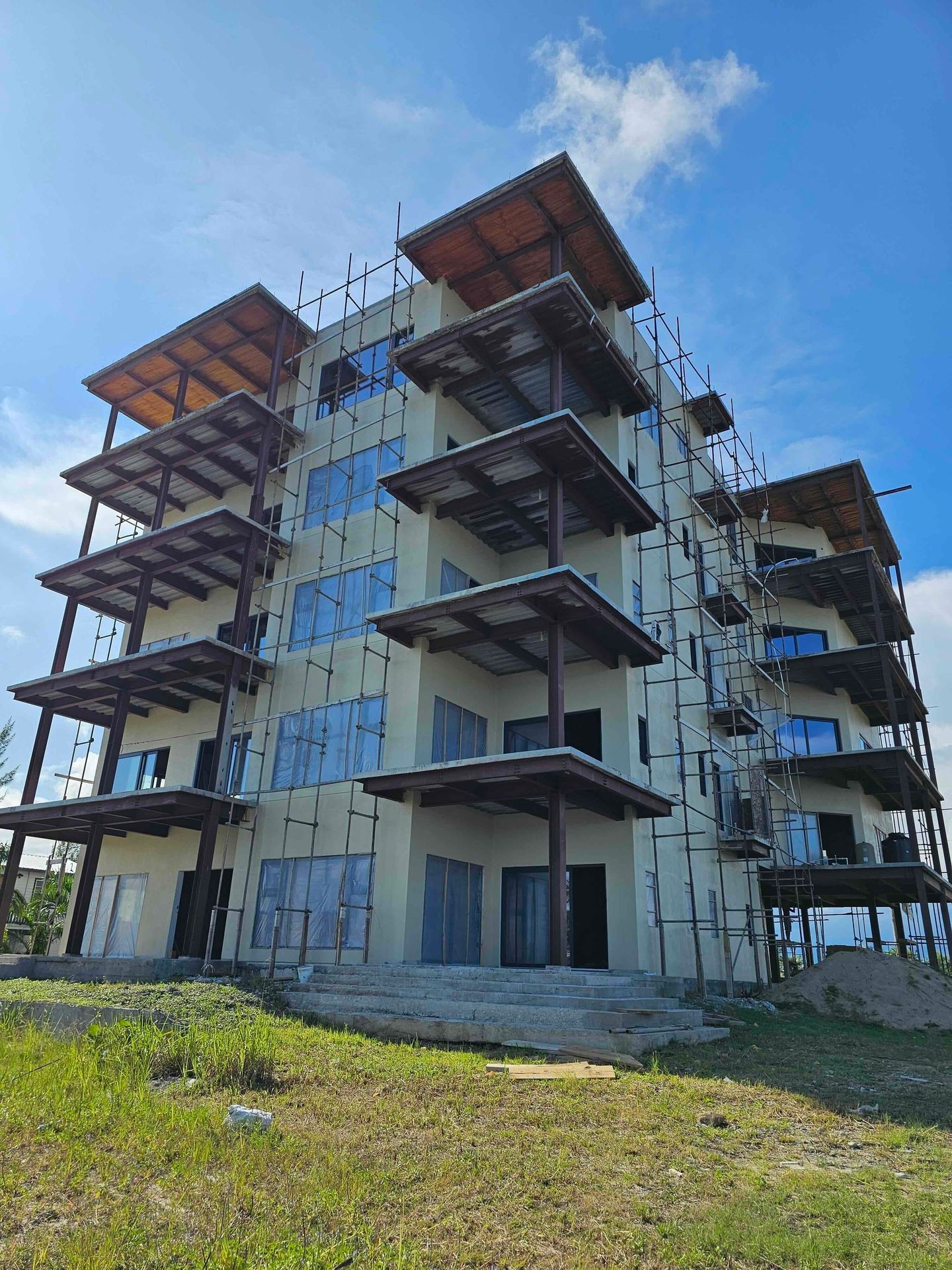 Four-story building under construction with scaffolding and unfinished balconies against a blue sky.